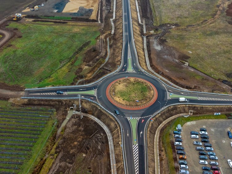 Vedere aeriană a unui drum aflat în construcție. În prim-plan se vede un sens giratoriu nou construită, iar în fundal un pod traversează o linie de cale ferată.