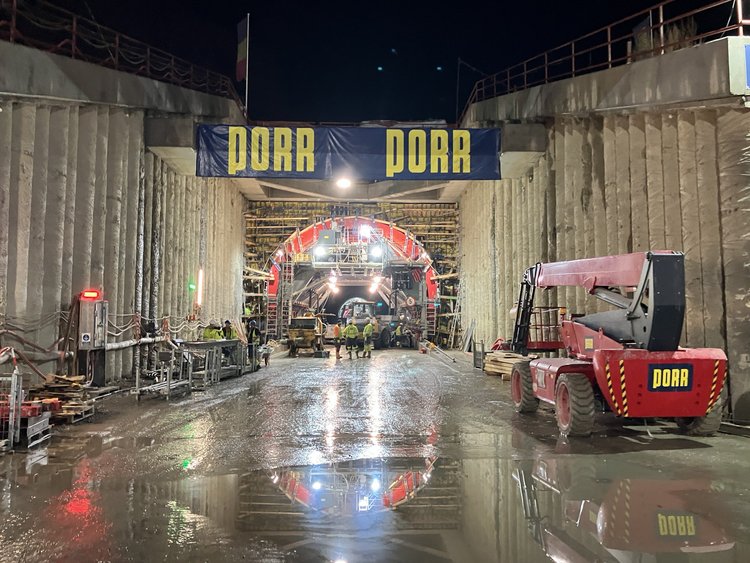 An illuminated tunnel construction site, with the tunnel entrance flanked by bored pile walls. Several people are working in front of it. On the right is a red lifting platform bearing the PORR logo; above the tunnel entrance are two large PORR banners.