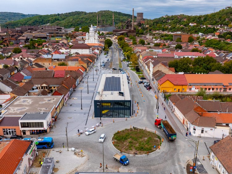 Vedere aeriană asupra unei stații moderne de tramvai, cu panouri fotovoltaice pe acoperiș. Zona este amenajată cu trotuare late și drumuri recent asfaltate.. În prim-plan, un sens giratoriu cu mai multe autovehicule angajate în trafic. Stația este înconjurată de locuințe de joasă înălțime, cu dealuri împădurite svizibile în zare,