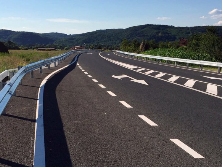 Photo: freshly asphalted and marked road, with a new pavement and guardrails, embedded in a rural landscape