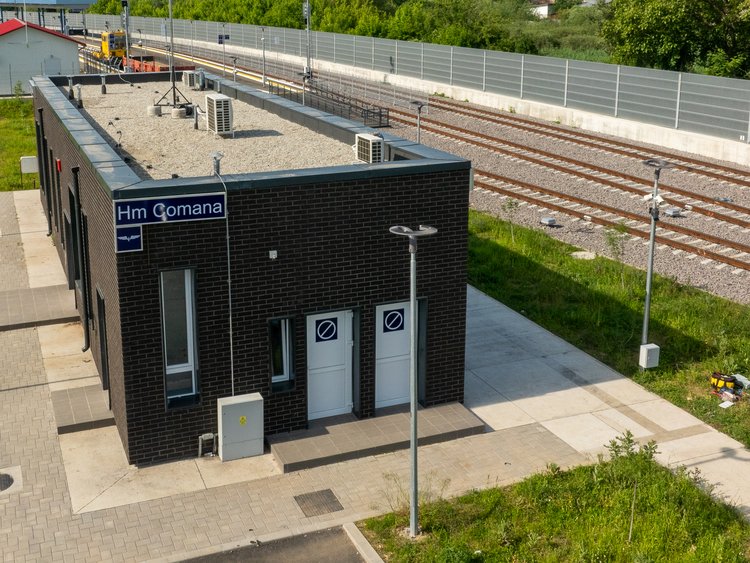 Peron modern, parțial acoperit, în centru, cu linii de cale ferată la stânga și dreapta sa. O zonă asfaltată lângă linia din stânga, iarbă în continuarea acesteia. Împrejmuire în fundal, pe partea dreaptă.