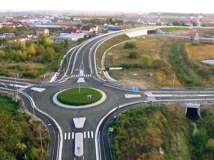 Foto: Luftaufnahme von vier Straßen, die in einem Kreisverkehr enden; die Überführung im Hintergrund; Umgebung von grünem Gras, Bäumen, einigen Häusern und geparkten Autos