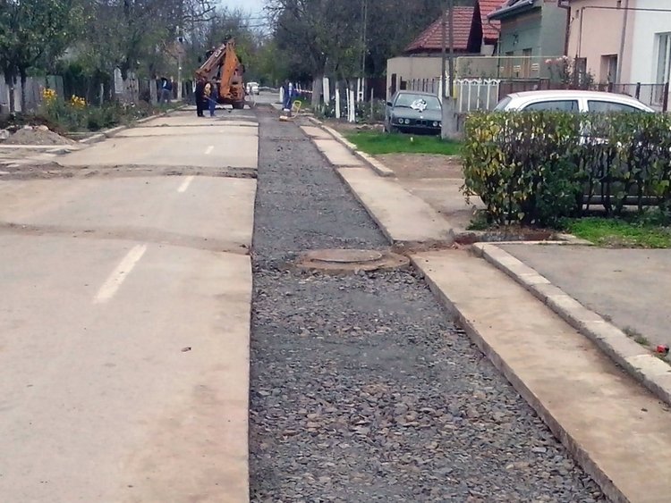 Photo: view of a street which has partly been opened and covered with porous asphalt; on the right, houses, driveways, cars, power lines; in the background, construction machines, trees, a mostly grey and cloudy sky